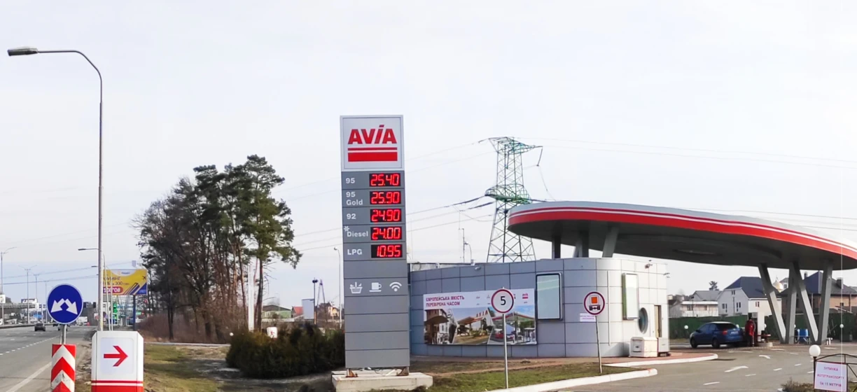Fuel prices and advertising signs at a city filling station with compact cars and automotive mirrors. Hard limit 125 characters: "City fuel station with compact cars, fuel dispenser, and advertising signs. Prices for gasoline, diesel, and LPG displayed."