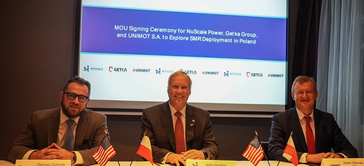Tłumaczenie: Profesjonaliści w garniturach podczas ceremonii podpisania memorandum o porozumieniu między NuScale Power, Get ka Group i UNIMOT S.A. w Polsce, z wystawionymi urządzeniami elektronicznymi i mikrofonami.